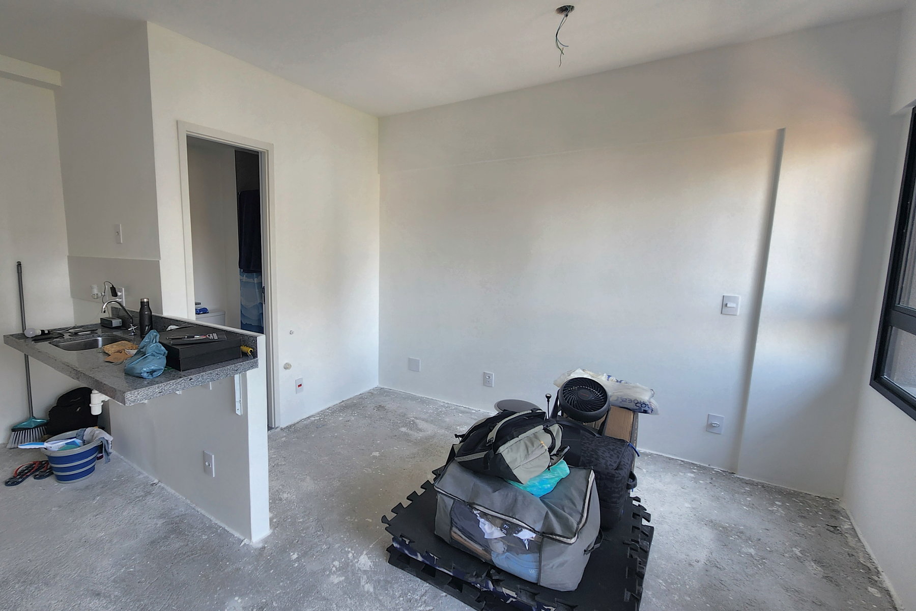 Studio de 26,36 m² como entregue pela construtora: paredes brancas, piso no contrapiso, bancada de apoio com cuba sobre mão-francesa, mureta baixa e porta do banheiro ao fundo