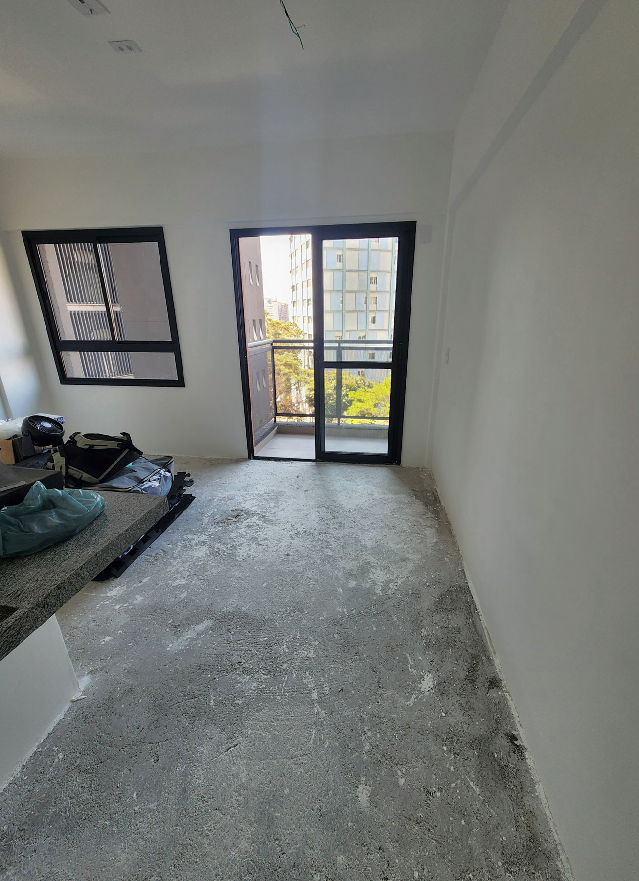 Studio vazio como entregue pela construtora, visto da cozinha: piso no contrapiso, fiação aparente no teto, janela e porta da varanda ao fundo