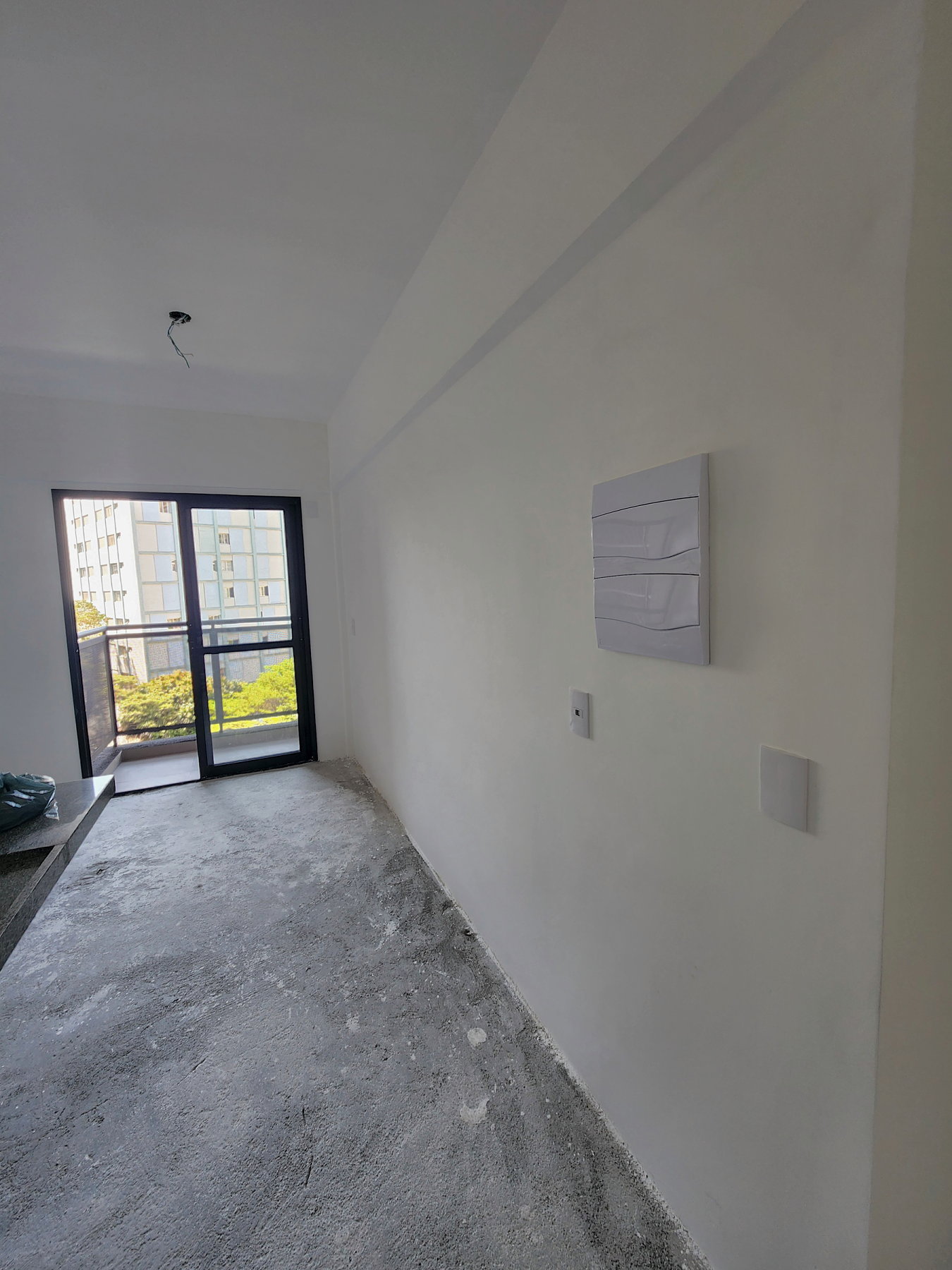Parede lateral do studio antes da reforma: parede branca lisa com o quadro de disjuntores e dois pontos elétricos, piso no contrapiso e a porta da varanda ao fundo
