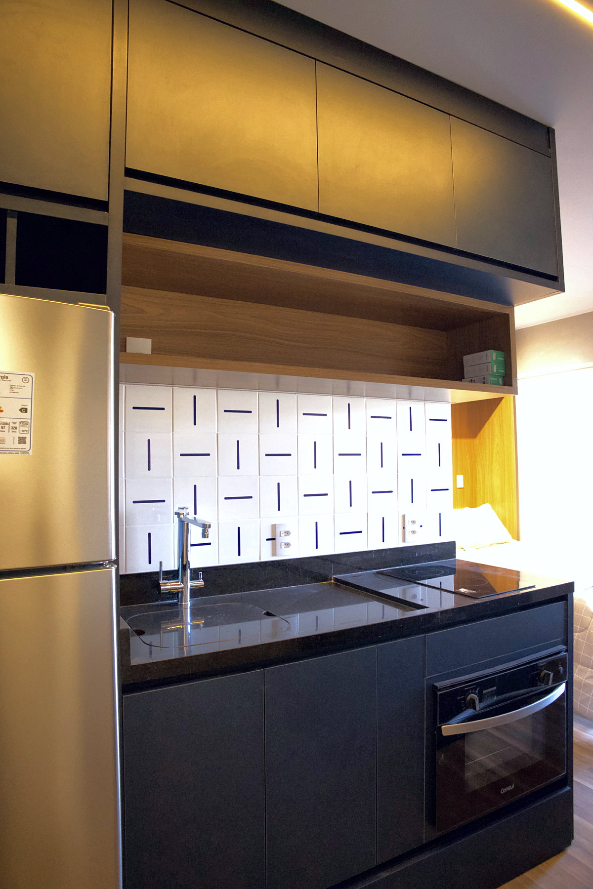 Cozinha pronta do studio: marcenaria azul-petróleo com torre da geladeira, prateleira em madeira, azulejos brancos com traço azul, bancada de granito preto com cooktop de indução e forno embutido
