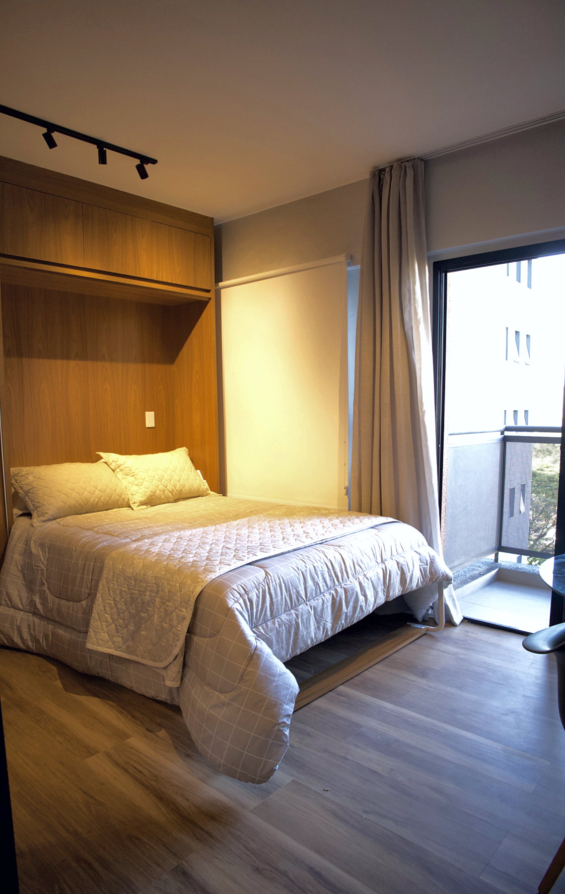 Cama de casal escamoteável aberta, saindo do armário de madeira clara, com a porta da varanda e a persiana ao lado, no studio pronto