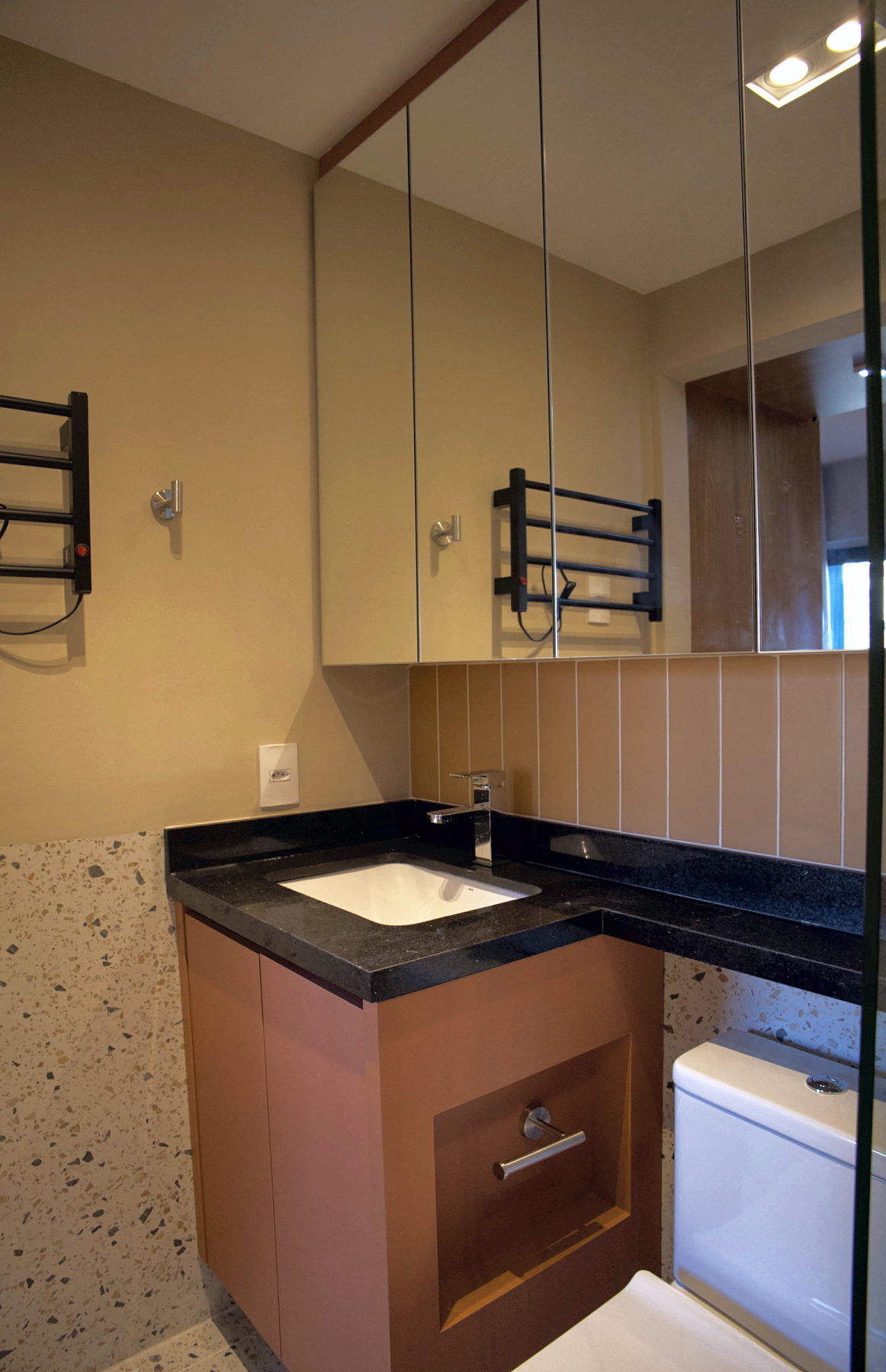 Bancada do banheiro em granito preto com cuba de embutir branca e torneira monocomando, gabinete terracota suspenso, espelheira, toalheiro elétrico preto e meia-parede em porcelanato Confete