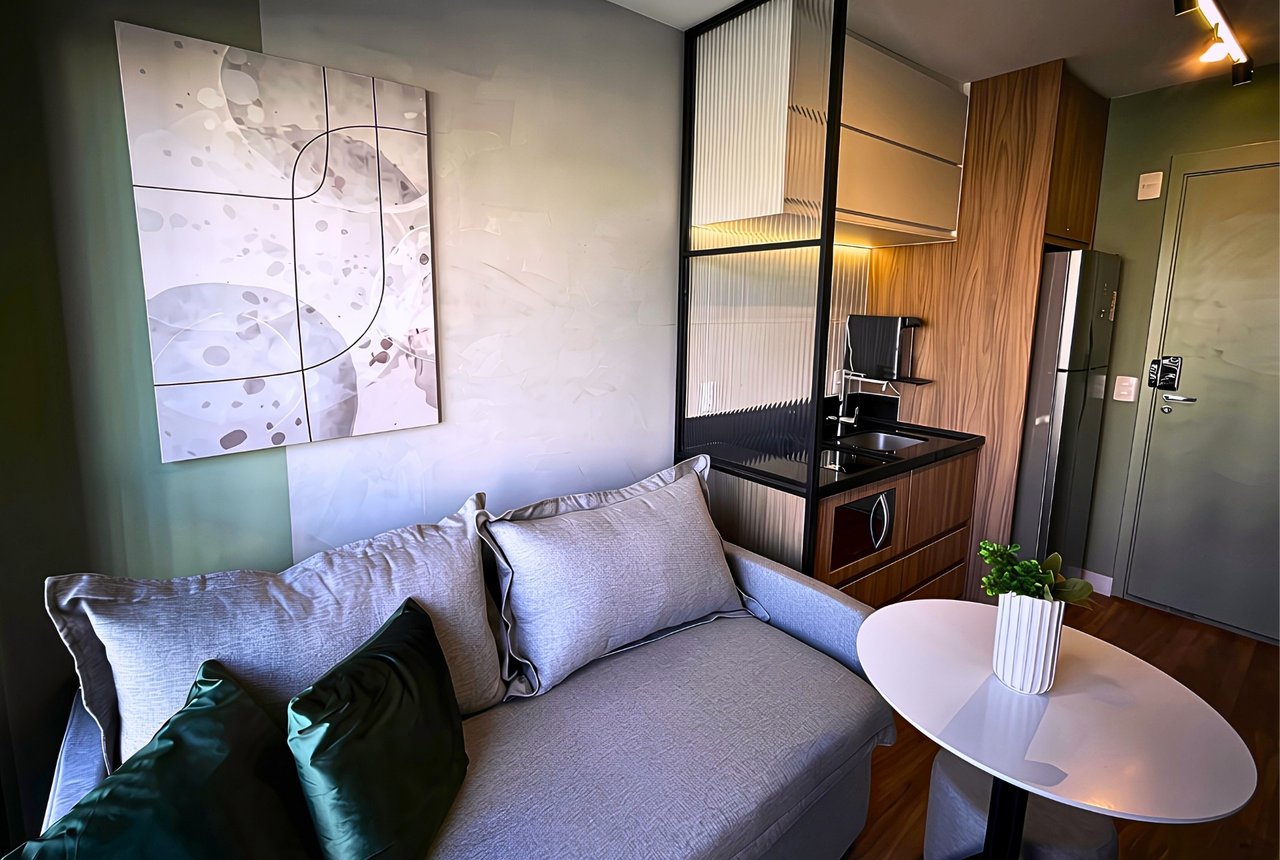 Studio com sofá cinza, almofadas verdes, cozinha planejada atrás de vidro e quadro de arte na parede